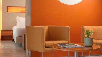 A modern hotel room with a bed, two orange armchairs, a small table with a magazine and a plant, and a wall with a circular light fixture.
