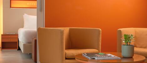 Una habitación de hotel moderna con una cama, dos sillones naranjas, una mesita con una revista y una planta, y una pared con una lámpara circular.
