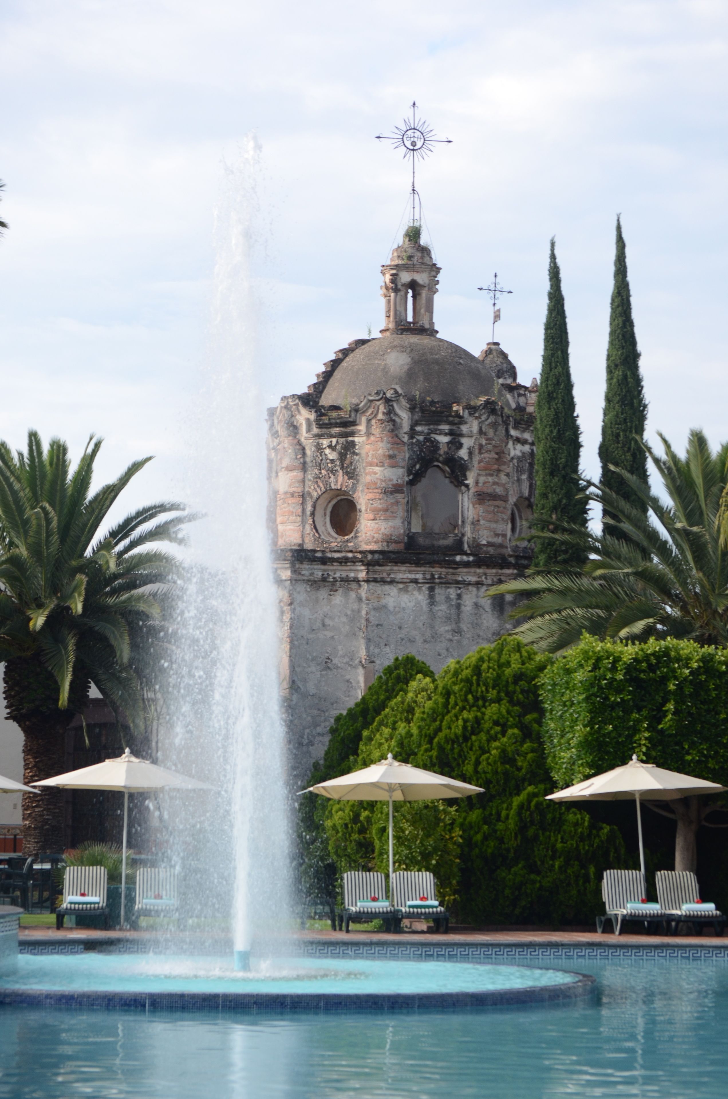 Una fuente frente a un edificio histórico con cúpula y aguja, rodeado de palmeras y sillones de descanso.