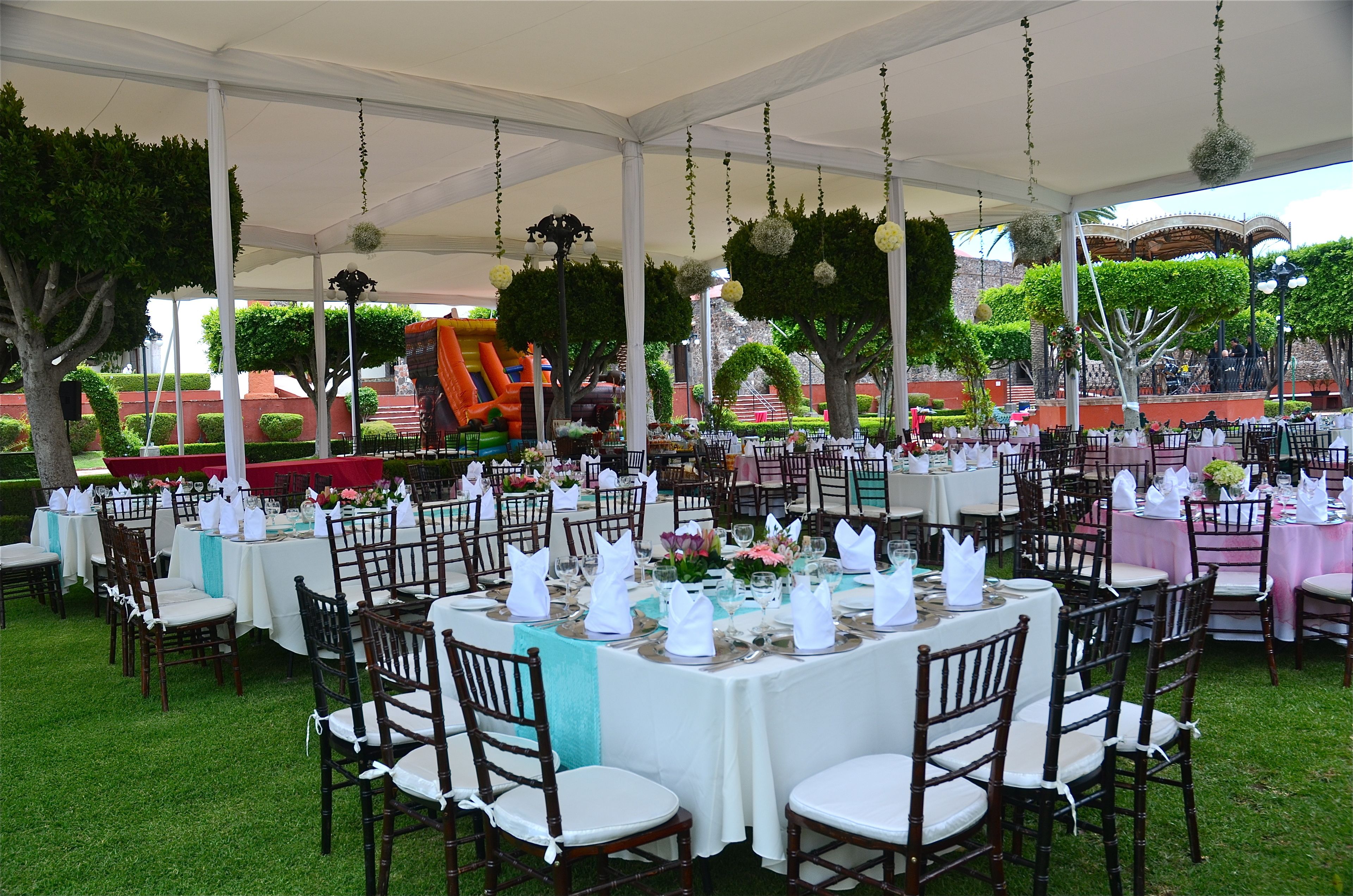 Un espacio para eventos al aire libre con mesas redondas dispuestas para cenar, rodeado de sillas, bajo una carpa con adornos colgantes.
