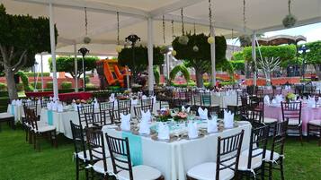Un espacio para eventos al aire libre con mesas redondas dispuestas para cenar, rodeado de sillas, bajo una carpa con adornos colgantes.