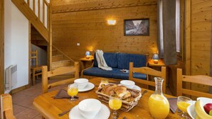 L’intérieur d’un chalet en bois présente une table à manger dressée pour le petit-déjeuner, avec des croissants, des fruits et du jus d’orange.
