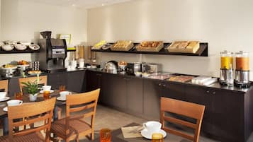 Un bufé de desayuno con diversas opciones de comida y bebidas, una máquina de café y mesas dispuestas.