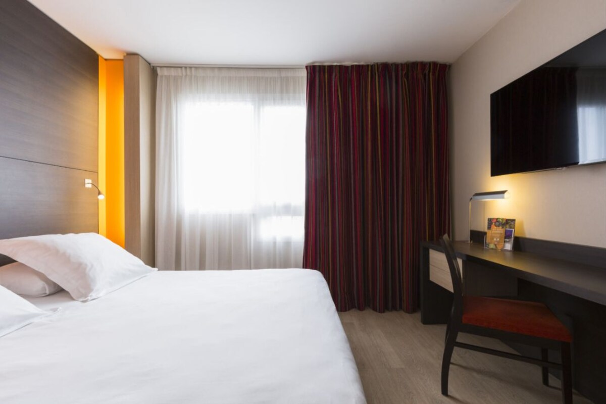Une chambre d’hôtel équipée d’un lit, d’un bureau, d’une chaise, d’une télévision et d’une fenêtre avec des rideaux.