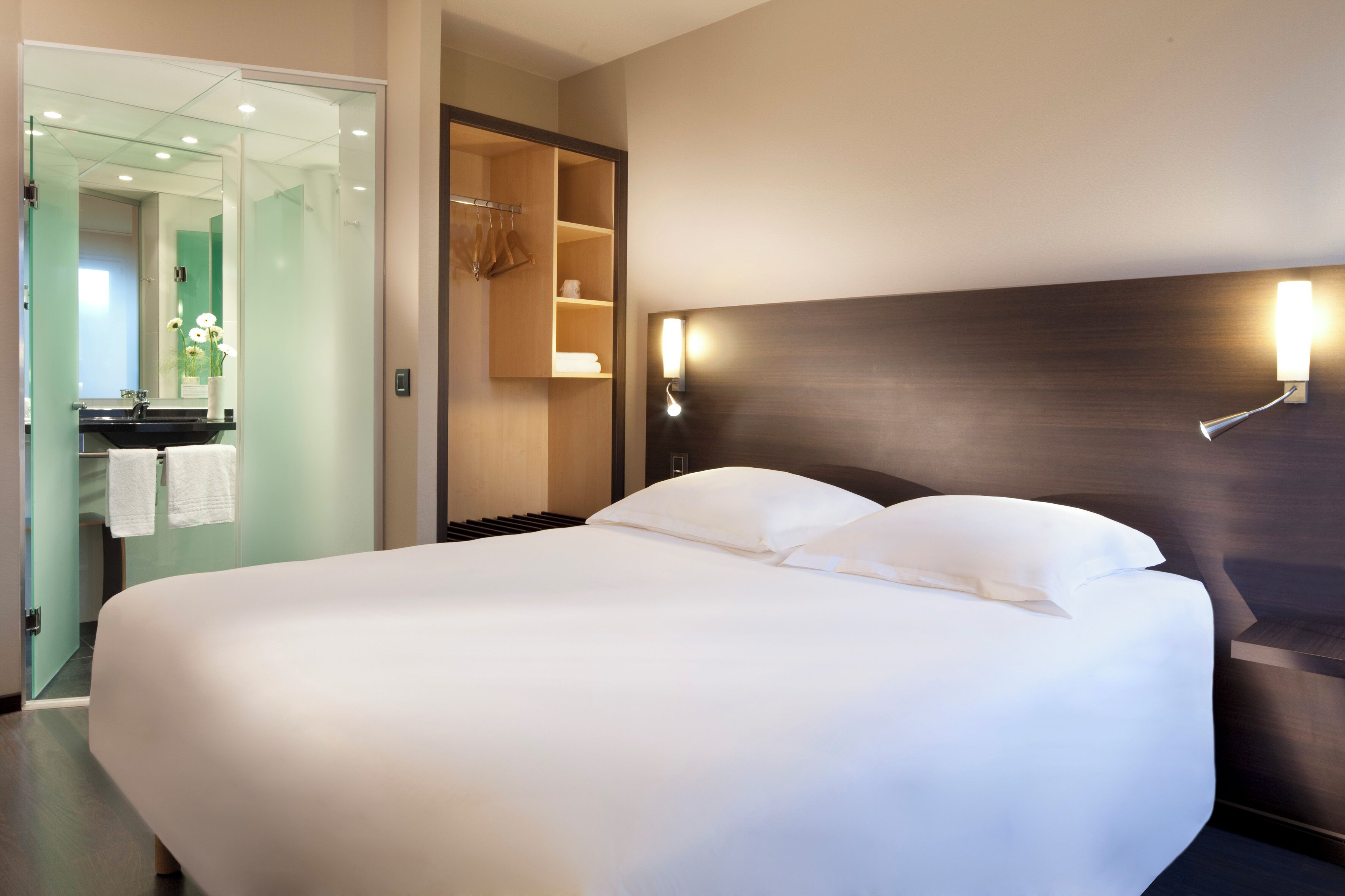 A hotel room with a large bed, a bathroom with a glass partition, and a wall-mounted light fixture.