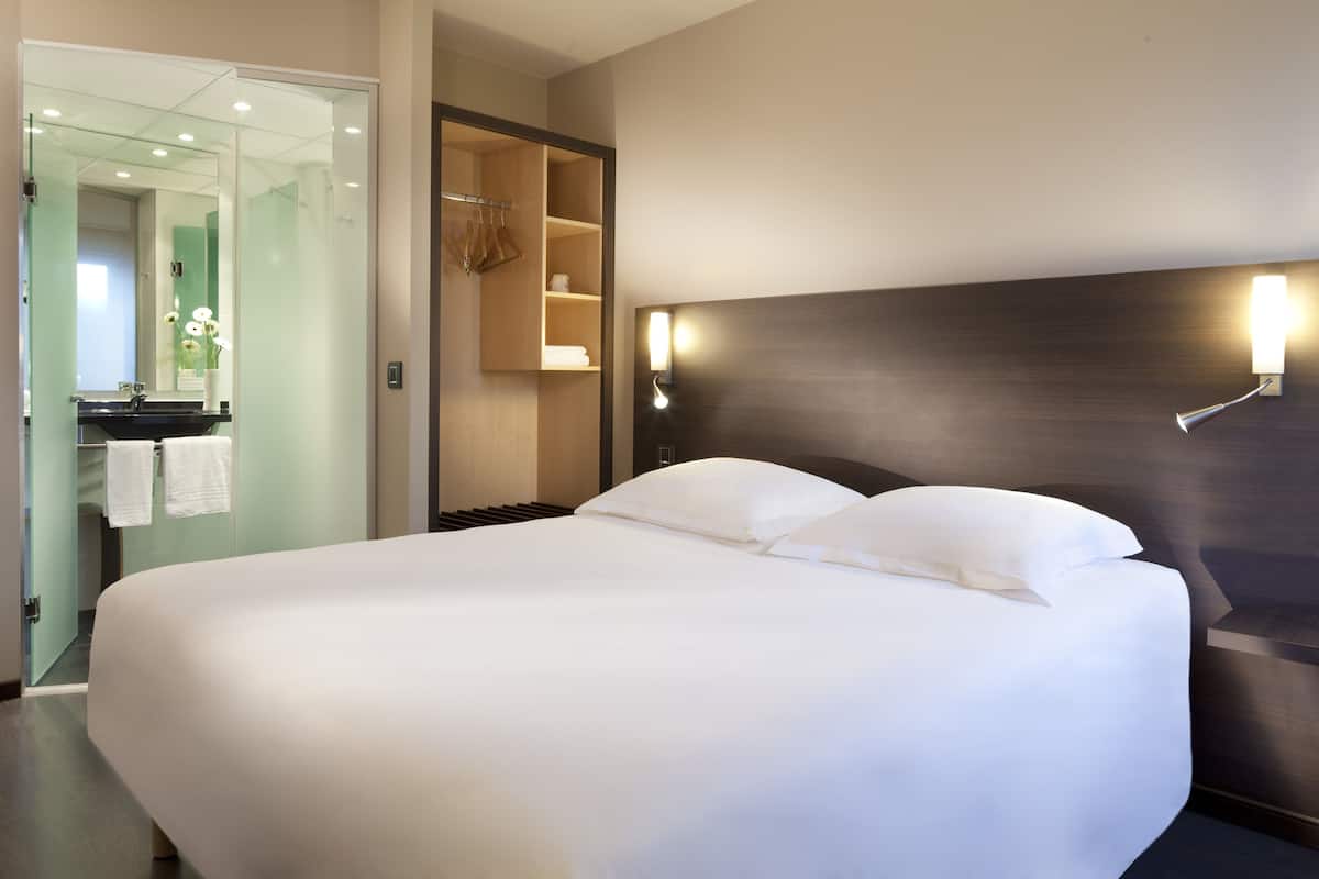 Una habitación de hotel moderna con una cama grande, cabina de ducha de vidrio, armario de madera y una lámpara fijada a la pared.