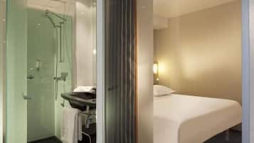 Habitación de hotel con ducha envuelta en vidrio, un lavamanos con toalla y una cama con almohada blanca.