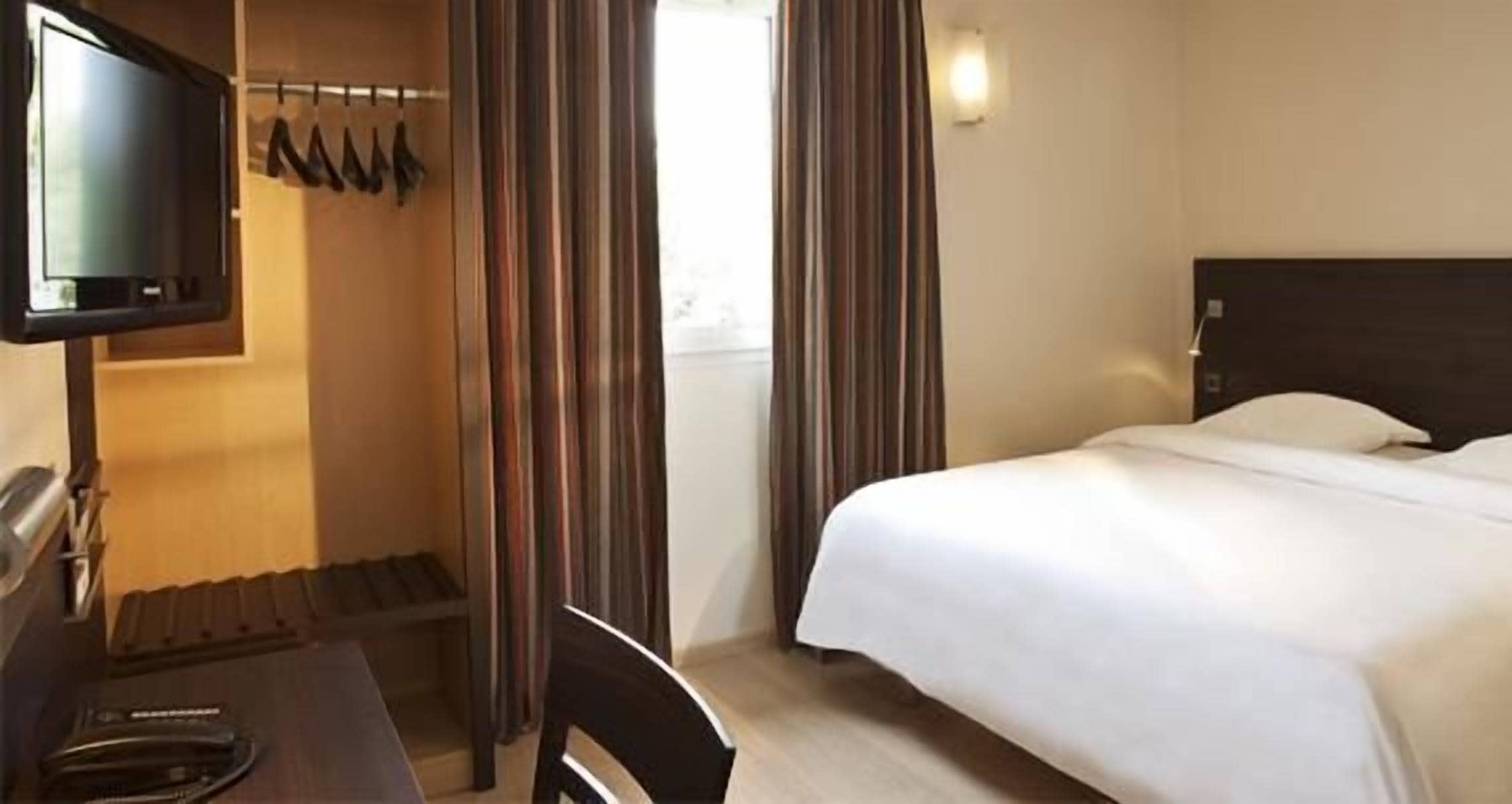 Habitación de hotel con cama, escritorio con silla, televisor y ventana con cortinas.