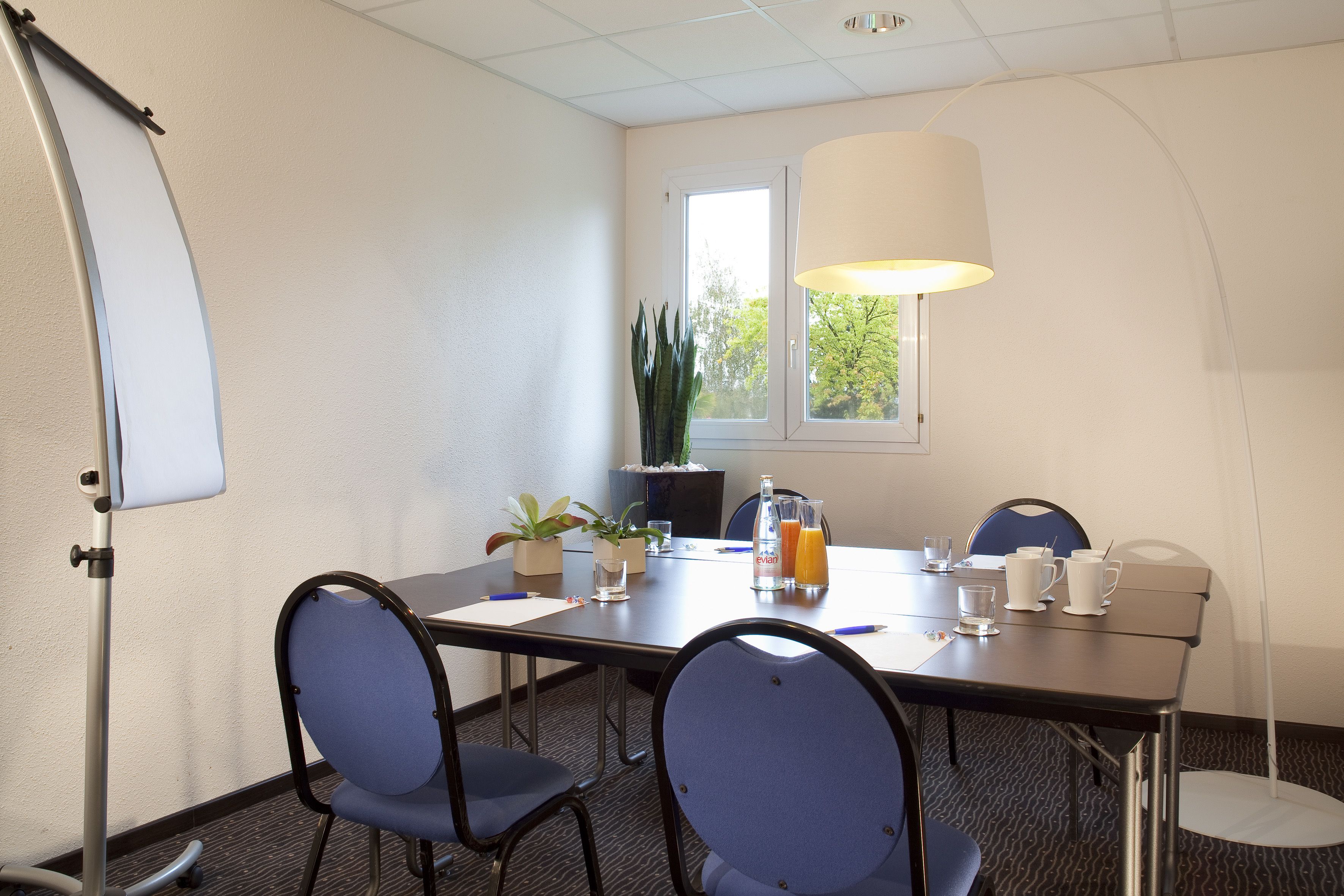 A conference room with a whiteboard, a table with chairs, a potted plant, a window with a view, and a floor lamp.
