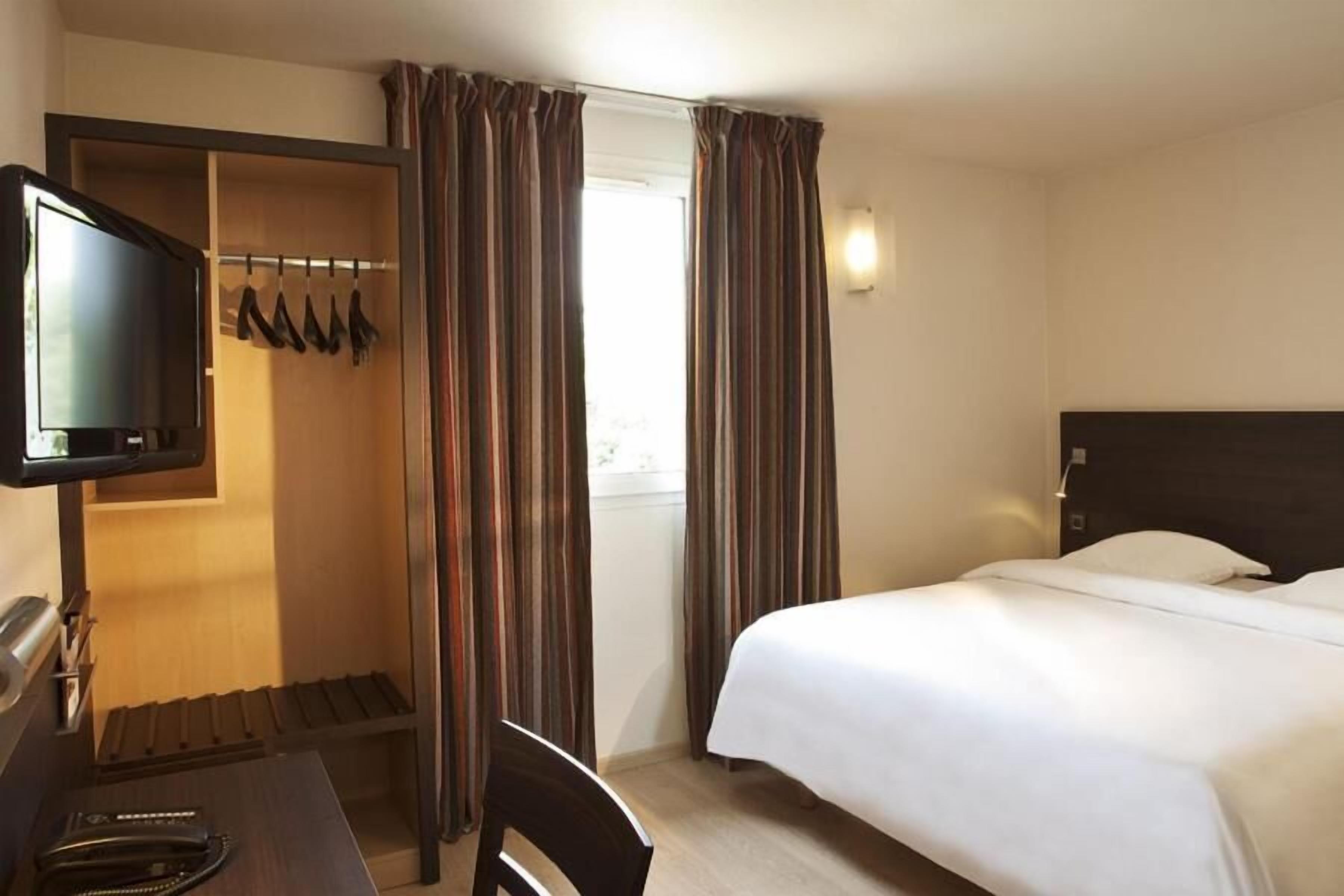 A hotel room with a bed, a desk with a television, a coat rack with shoes, and curtains.