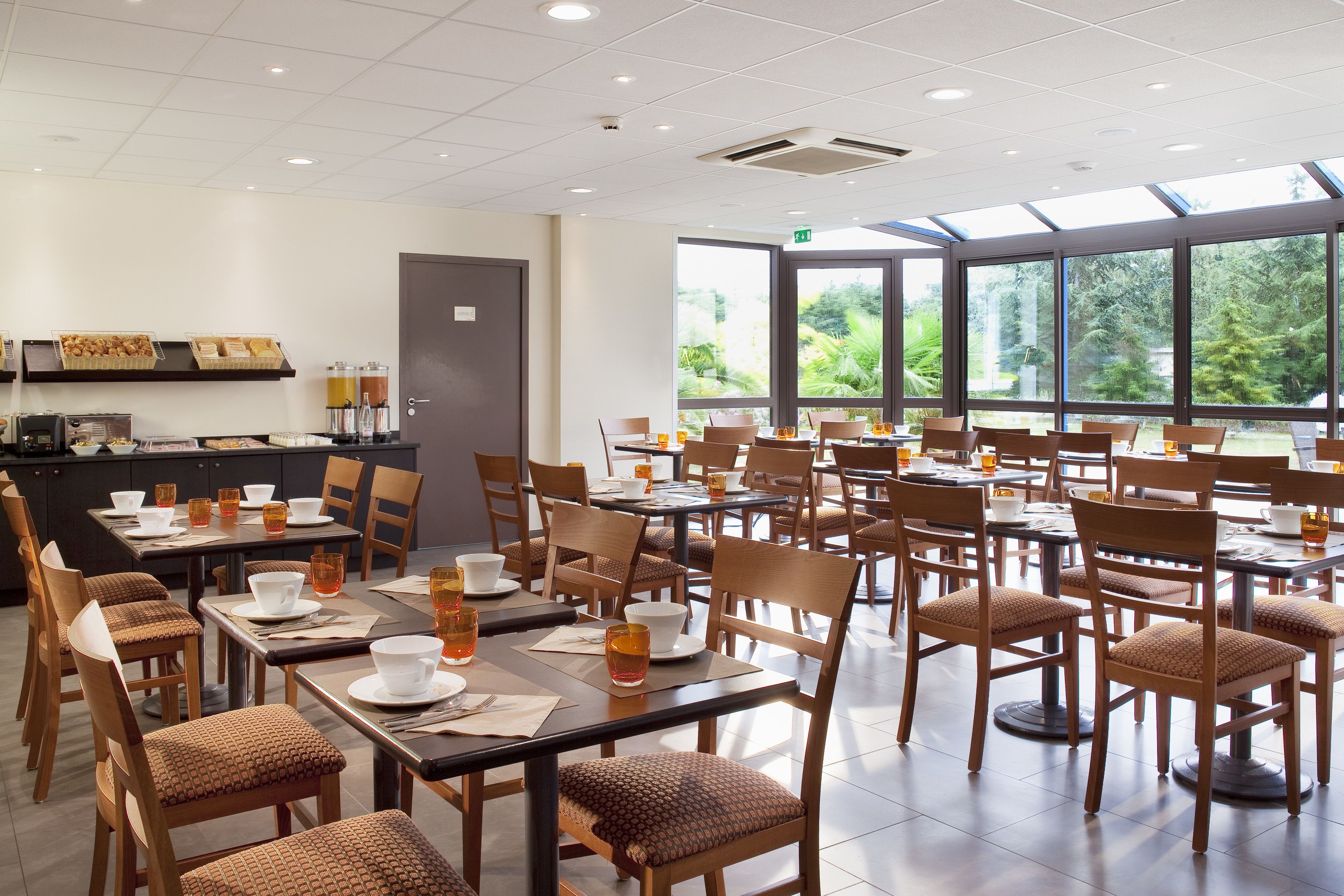 A spacious dining area with multiple tables and chairs, a counter with food items, and large windows offering a view of greenery.