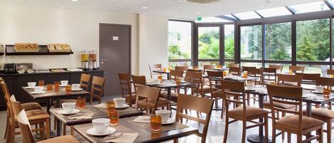 A spacious dining area with multiple tables and chairs, a counter with food items, and large windows offering a view of greenery.