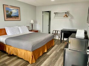A hotel room with a bed, a desk with a chair, a refrigerator, a lamp, and a painting on the wall. - The L Motel - Downtown Flagstaff (Flagstaff)