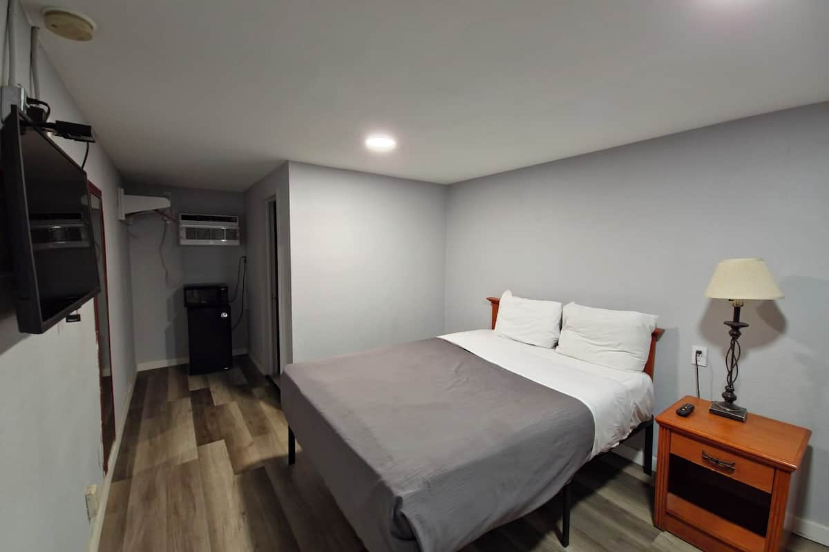 Quarto com cama, mesa de cabeceira, abajur e TV fixada na parede.