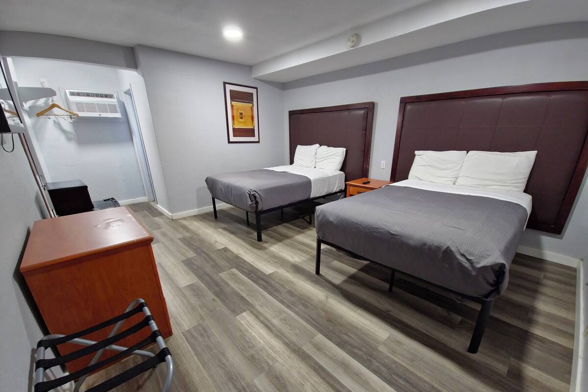 Quarto de hotel com duas camas de solteiro, uma cama de casal, cômoda de madeira, uma cadeira, escrivaninha e ar-condicionado fixado na parede.