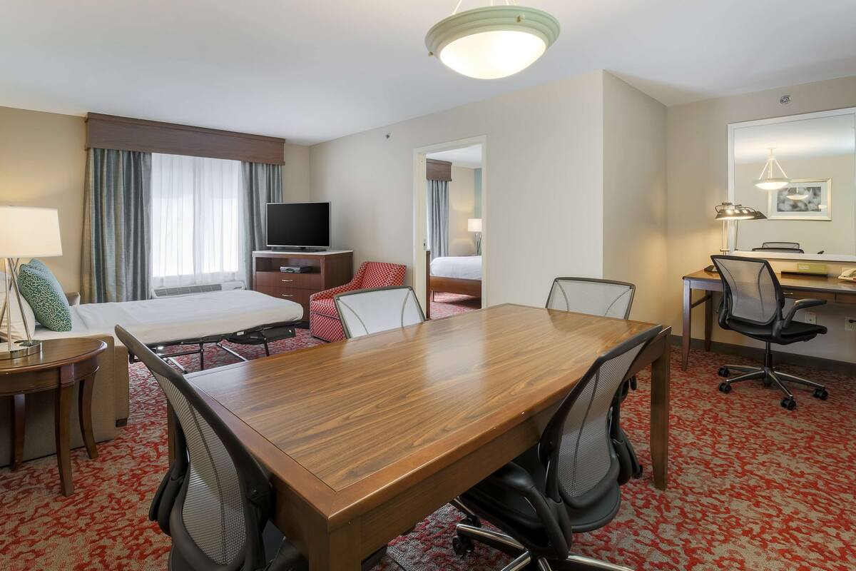 Hotellihuone, jossa on puinen kokouspöytä, kaksi tuolia, televisio, sänky, työpöytä tuolilla sekä suuri peili.