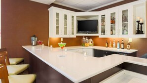 A bar area with a white countertop, bar stools, a television, and various bottles. - Hilton Garden Inn Nanuet (Nanuet)
