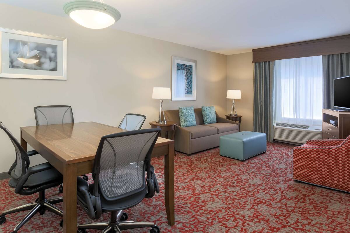 Hotellihuone, jossa on puinen pöytä, neljä toimistotuolia, kahdella tyynyllä varustettu sohva, punakuvioinen matto, televisio ja seinällä oleva kehystetty taideteos.