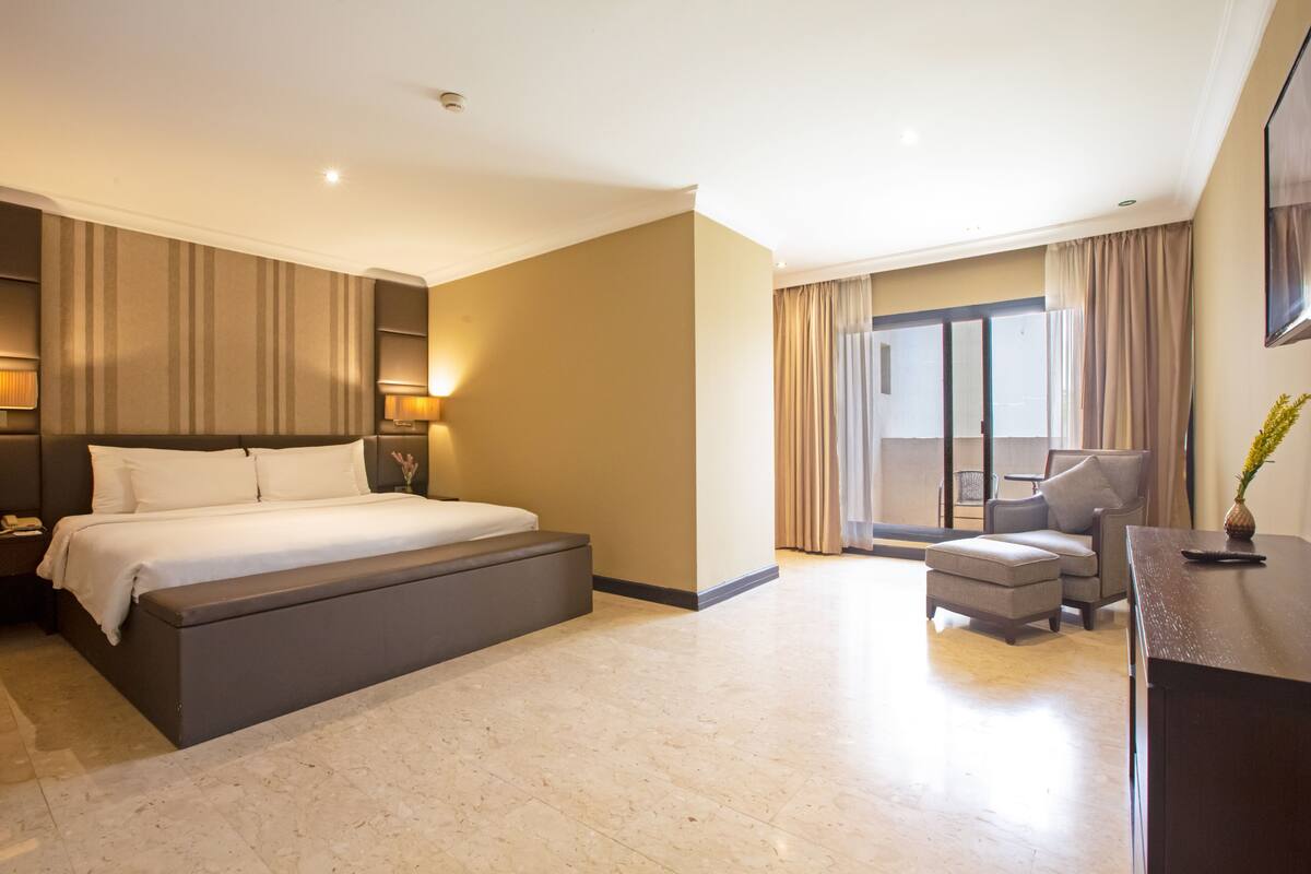 Une chambre d’hôtel moderne dotée d’un grand lit, d’un coin salon avec un canapé et un fauteuil, d’un bureau et offrant une vue sur l’extérieur grâce à une porte coulissante.