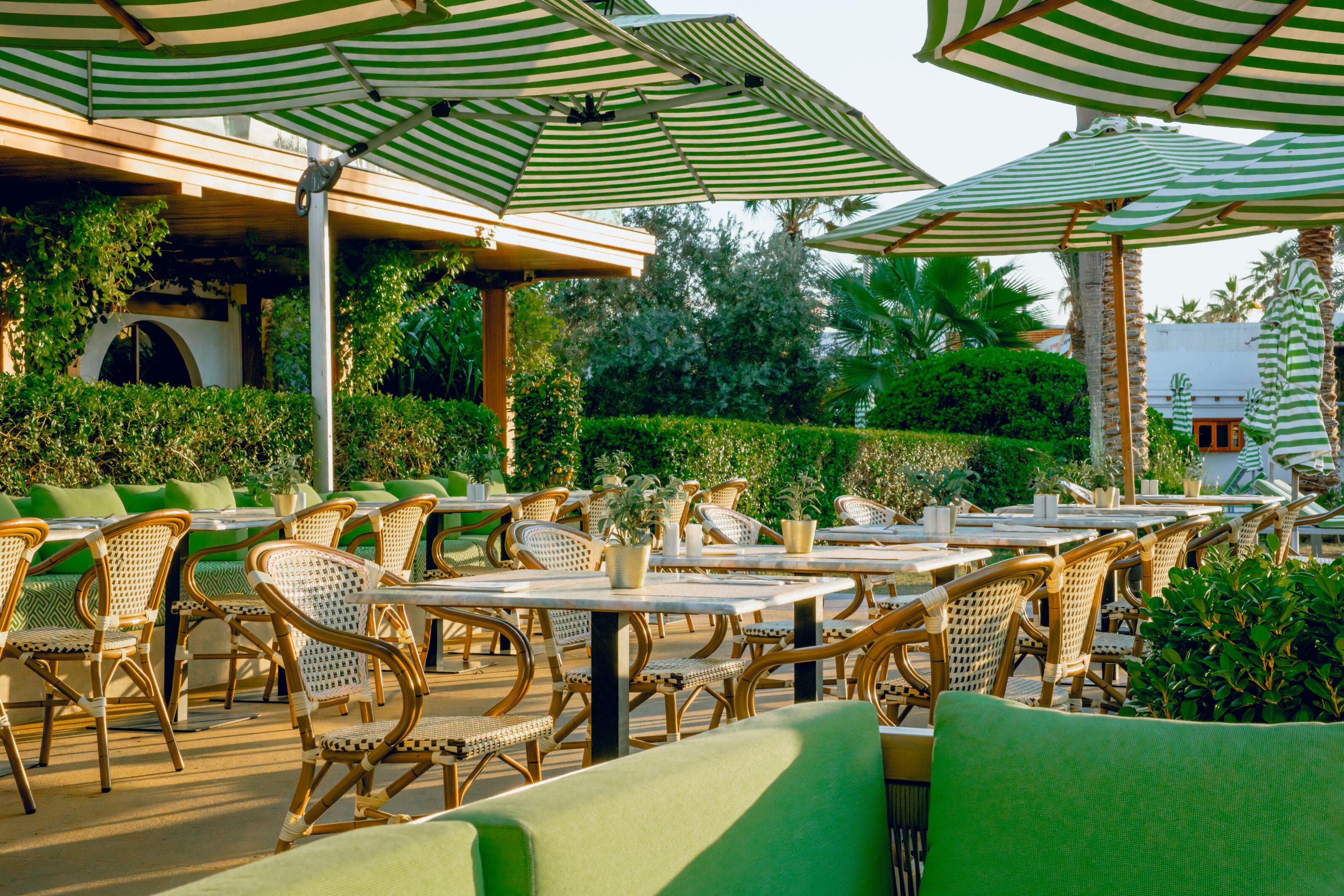 An outdoor seating area with wicker chairs and tables, green striped umbrellas, and lush greenery in the background.