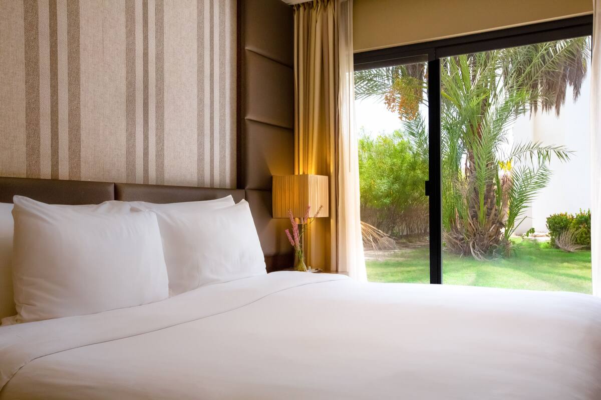 Une chambre d’hôtel avec un grand lit, une fenêtre donnant sur un paysage verdoyant et une lampe sur la table de chevet.