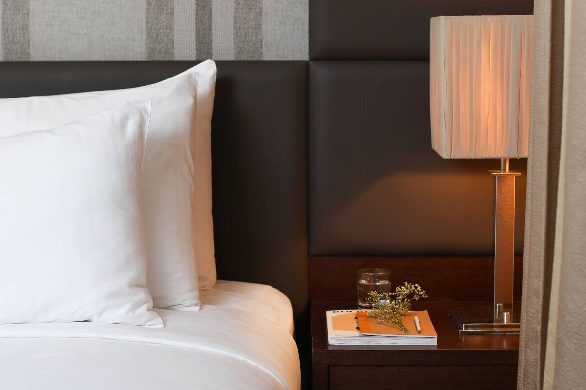 Un lit bien fait, avec du linge de lit blanc et des oreillers, une table de chevet avec une lampe, un livre et un verre d’eau.