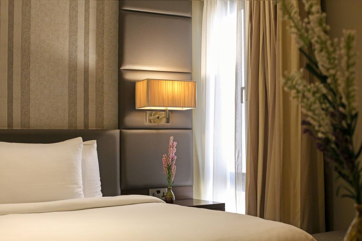 Une chambre d’hôtel avec un grand lit, une table de chevet avec une lampe, un vase de fleurs et une vue sur l’extérieur par la fenêtre.