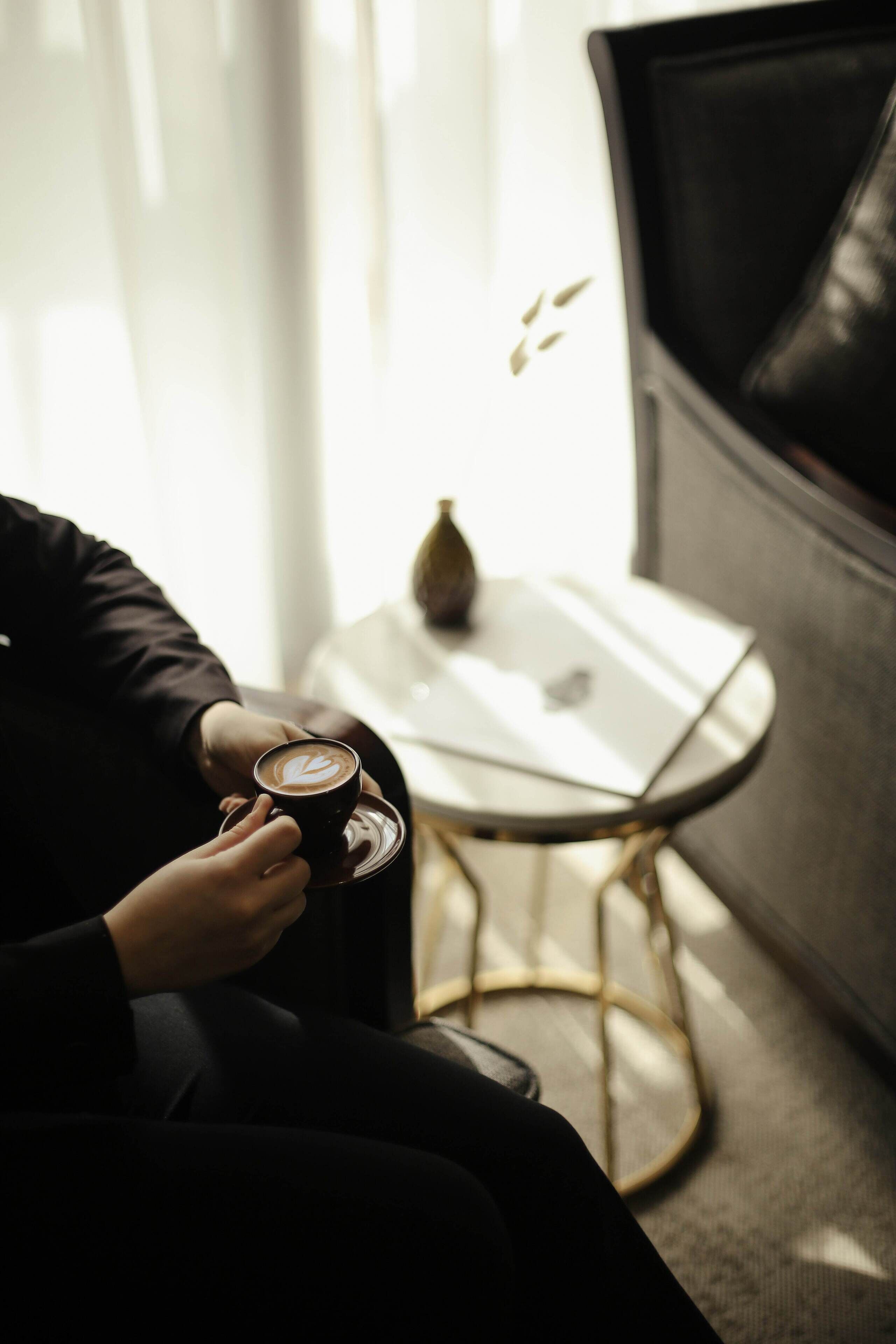 Una persona sostiene una taza de café en una habitación moderna con una pequeña mesa y un jarrón decorativo.