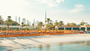 A beachside hotel with rows of orange sun loungers and umbrellas, a sandy beach, and a city skyline in the background. - Dubai Marine Beach Resort & Spa (Dubai)