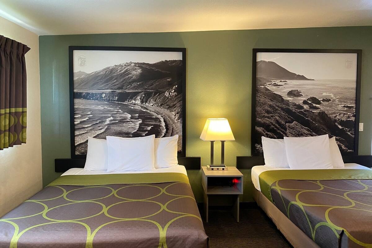 A hotel room with two beds, a nightstand with a lamp, and large landscape pictures on the walls.