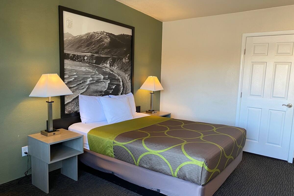 A hotel room with a bed, two bedside tables with lamps, a large framed picture on the wall, and a white door.