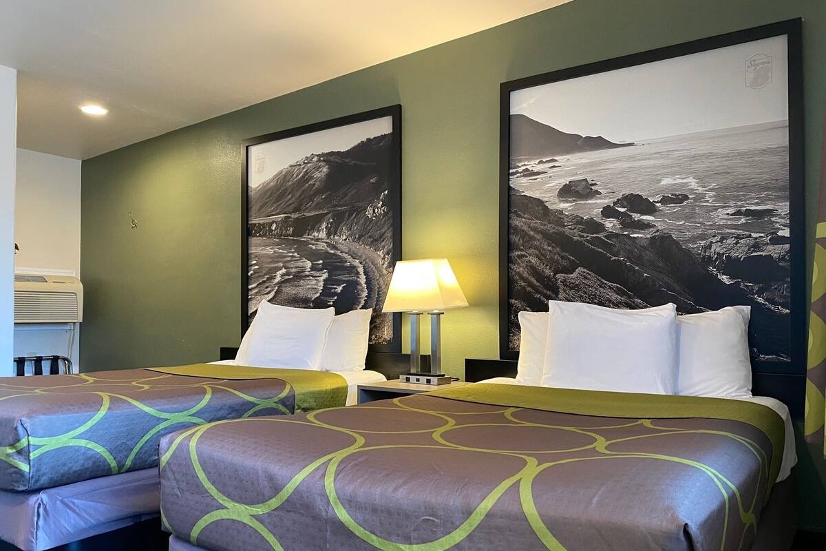A hotel room with two beds, a nightstand, a lamp, and two large landscape posters on the wall.