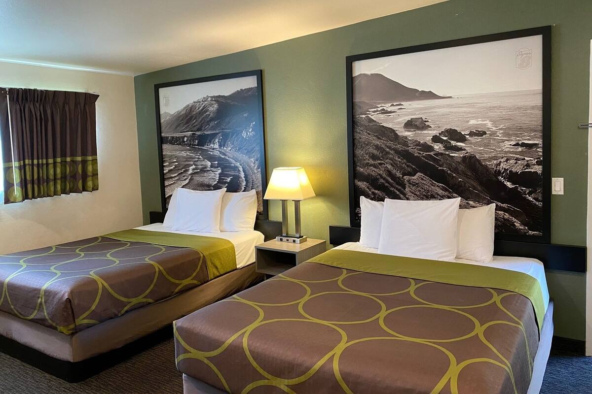 A hotel room with two beds, a nightstand with a lamp, and large landscape pictures on the walls.
