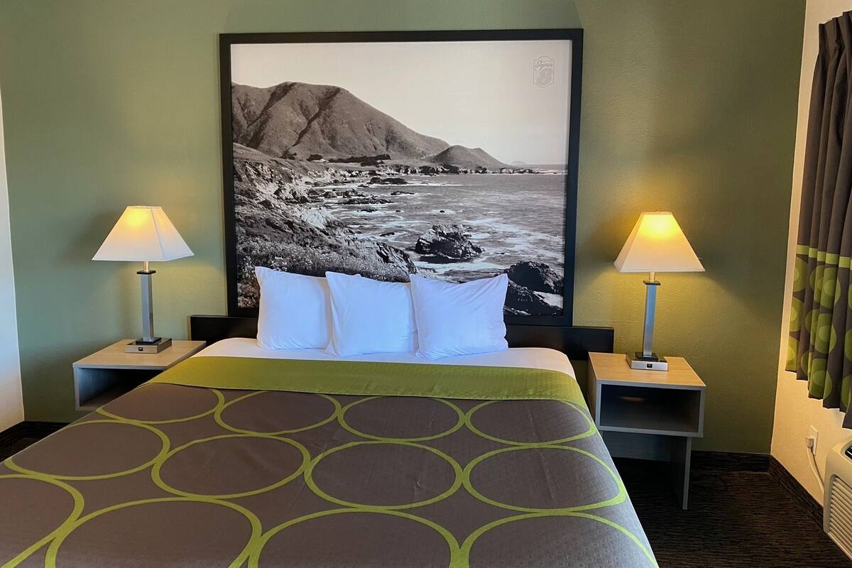 A hotel room with a bed, two bedside lamps, a large framed photograph of a coastal landscape, and a green patterned bedspread.