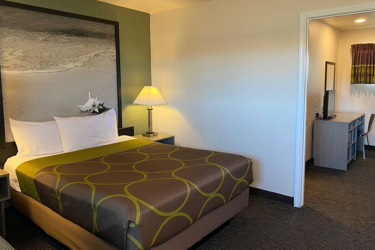 A hotel room with a bed, a desk, a lamp, and a painting of a beach scene.