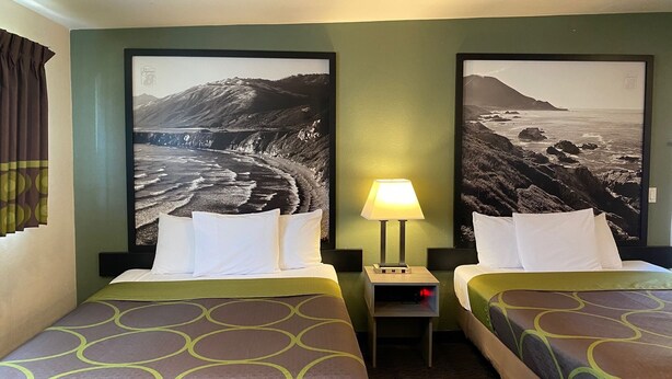 Ein Hotelzimmer mit zwei Betten, einem Nachttisch mit Lampe sowie zwei großformatigen Landschaftsbildern an den Wänden.