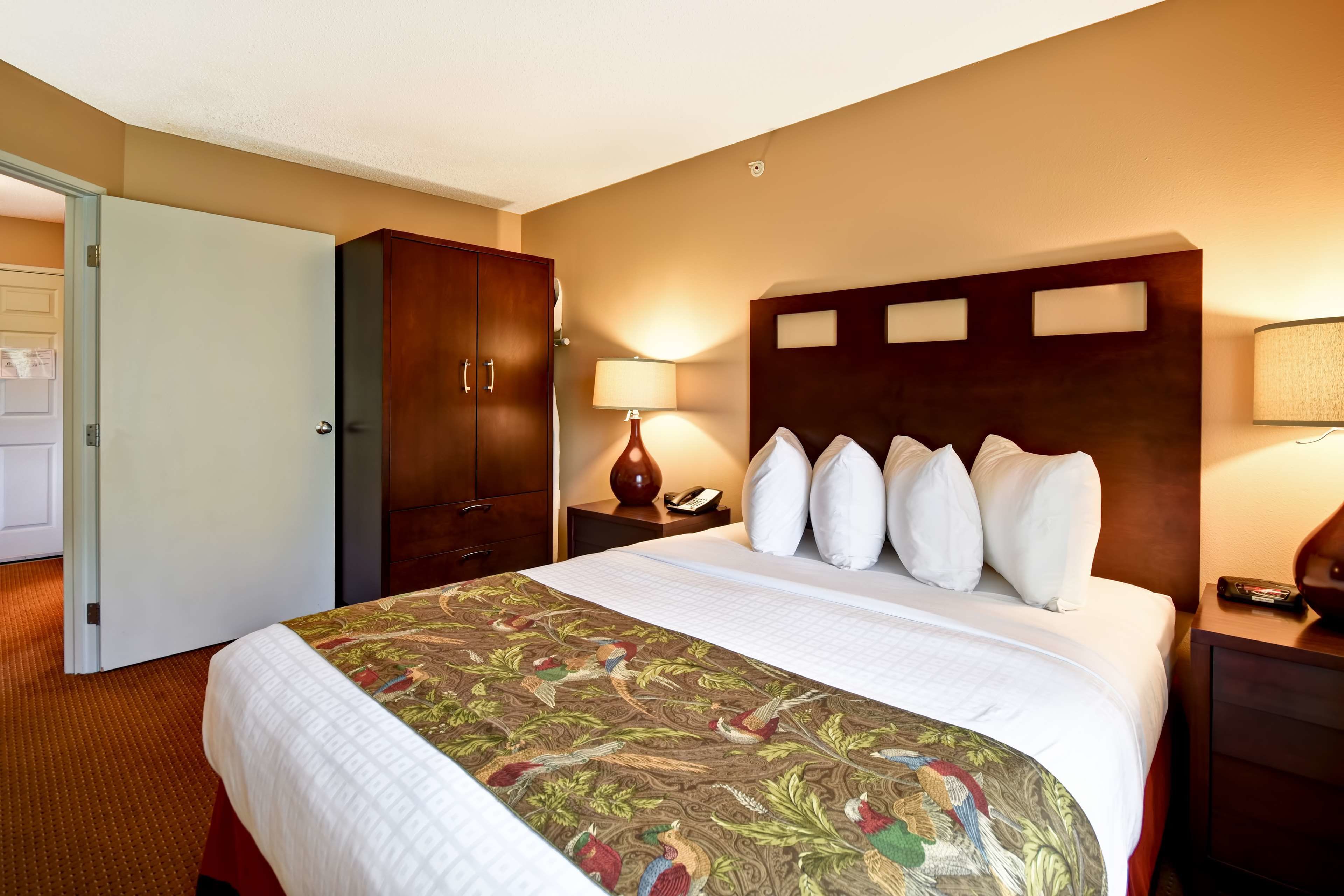 A hotel room with a large bed, a wooden wardrobe, a nightstand with a lamp, and a door leading to another room.