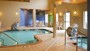 An indoor swimming pool with clear depth markings, a lifeguard warning sign, and a covered seating area with chairs. - GrandStay Hotel & Suites (La Crosse)