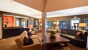 A spacious living room with a large sofa, a coffee table, and a fireplace. There is a reception area with a counter and a plant. - GrandStay Hotel & Suites (La Crosse)