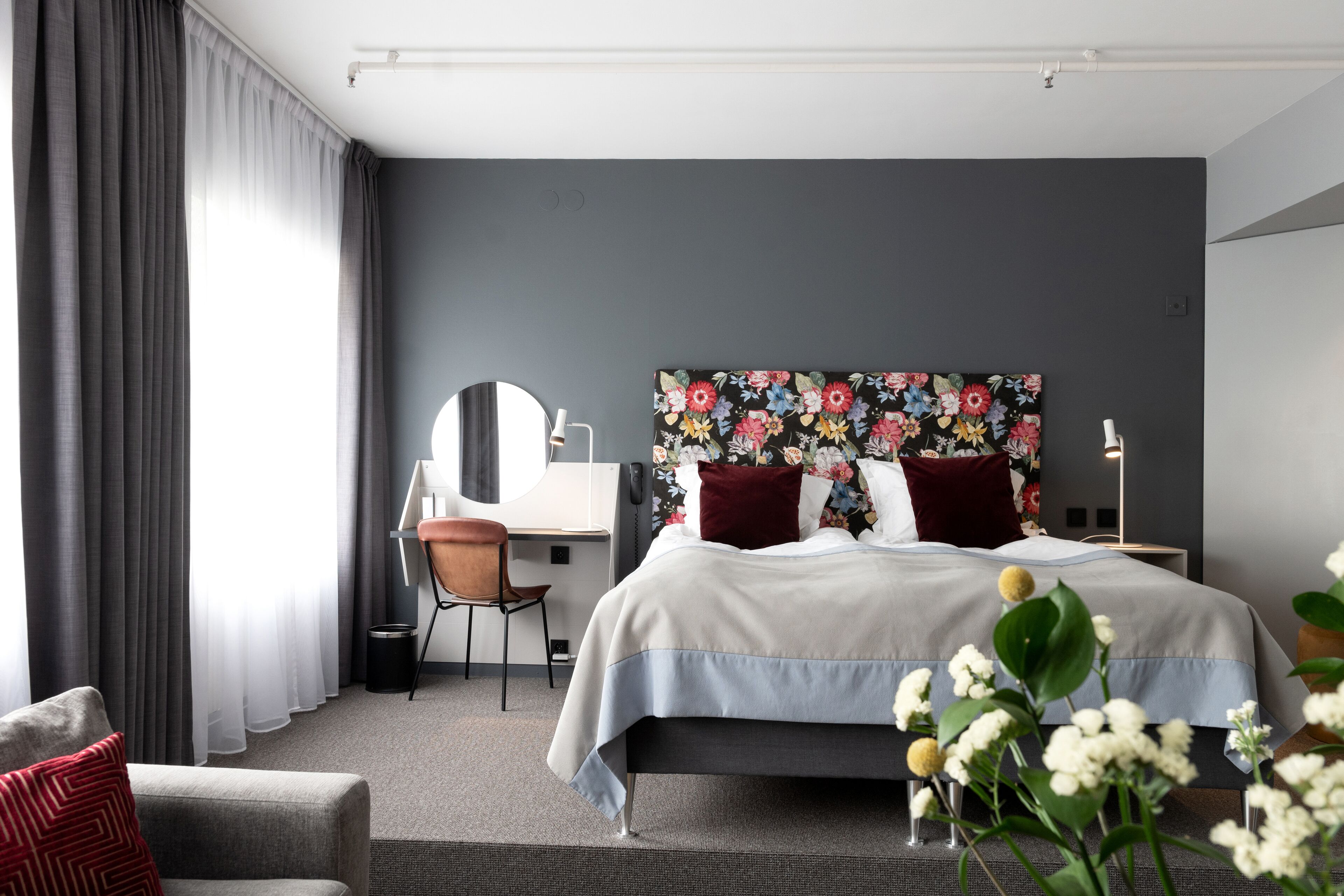 A modern hotel room with a large bed, a desk with a chair, a round mirror, and a floral headboard.