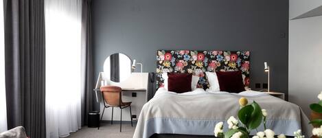 A modern hotel room with a large bed, a desk with a chair, a round mirror, and a floral headboard.
