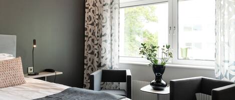 A hotel room with a bed, a desk, a chair, a potted plant, and a window with curtains.