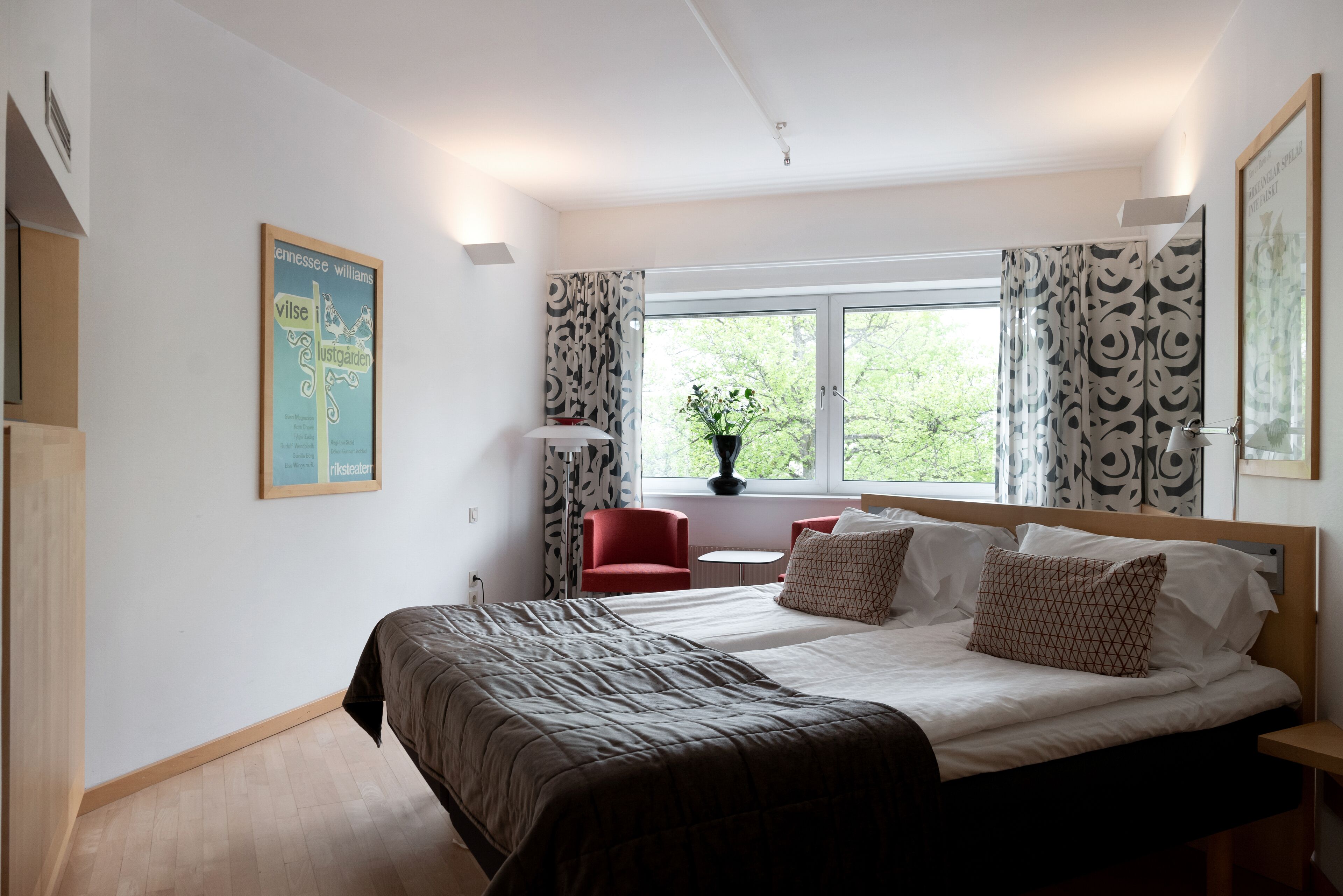 Une chambre d’hôtel moderne avec un grand lit, une petite table, une chaise rouge et une fenêtre avec des rideaux.