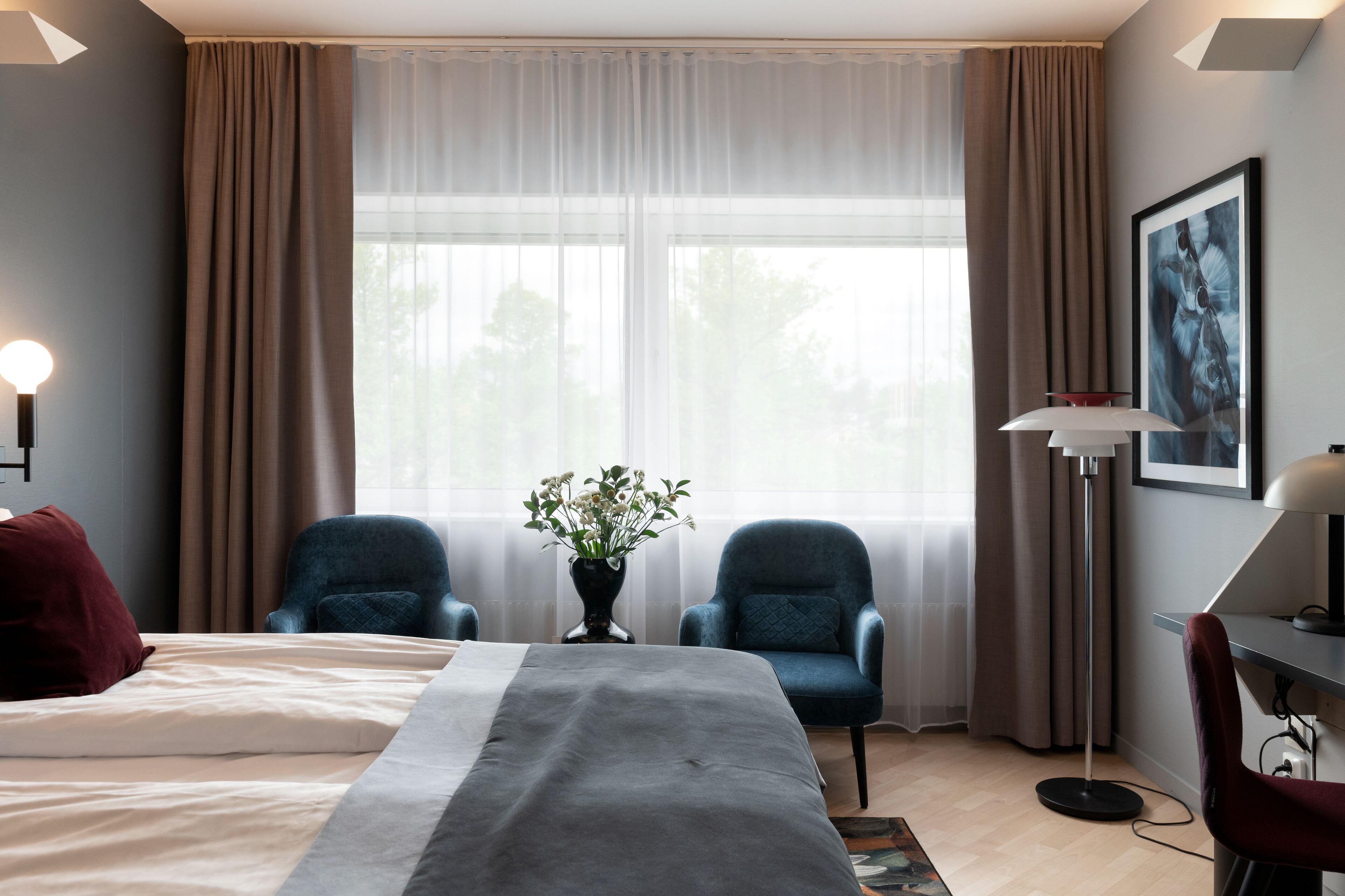 Een hotelkamer met een bed, twee fauteuils, een bureau, een lamp, een vaas met bloemen en een groot raam met gordijnen.