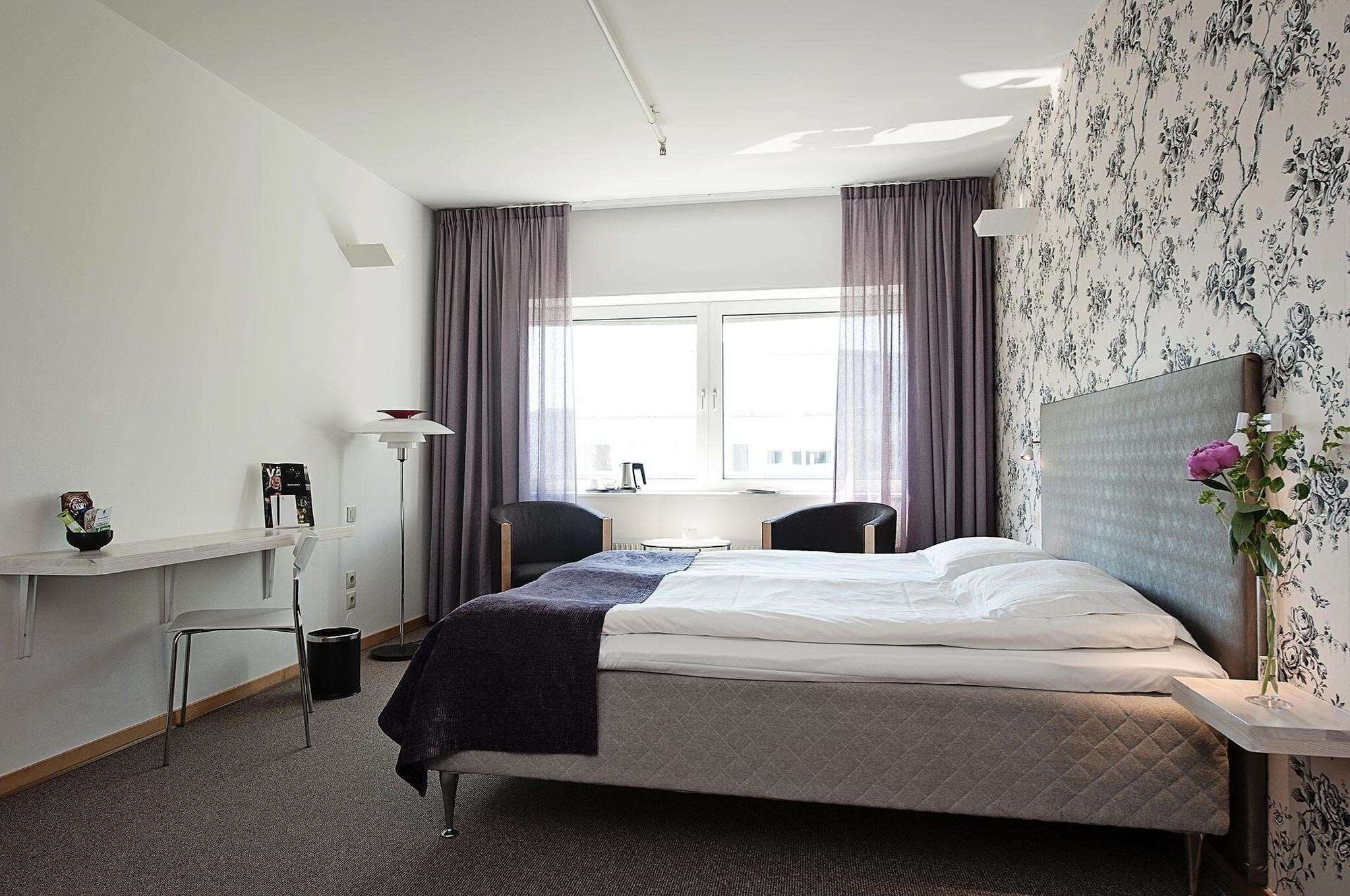 Quarto de hotel com cama, escrivaninha, cadeira, mesa pequena e janela com cortinas.