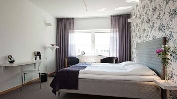 Quarto de hotel com cama, escrivaninha, cadeira, mesa pequena e janela com cortinas.