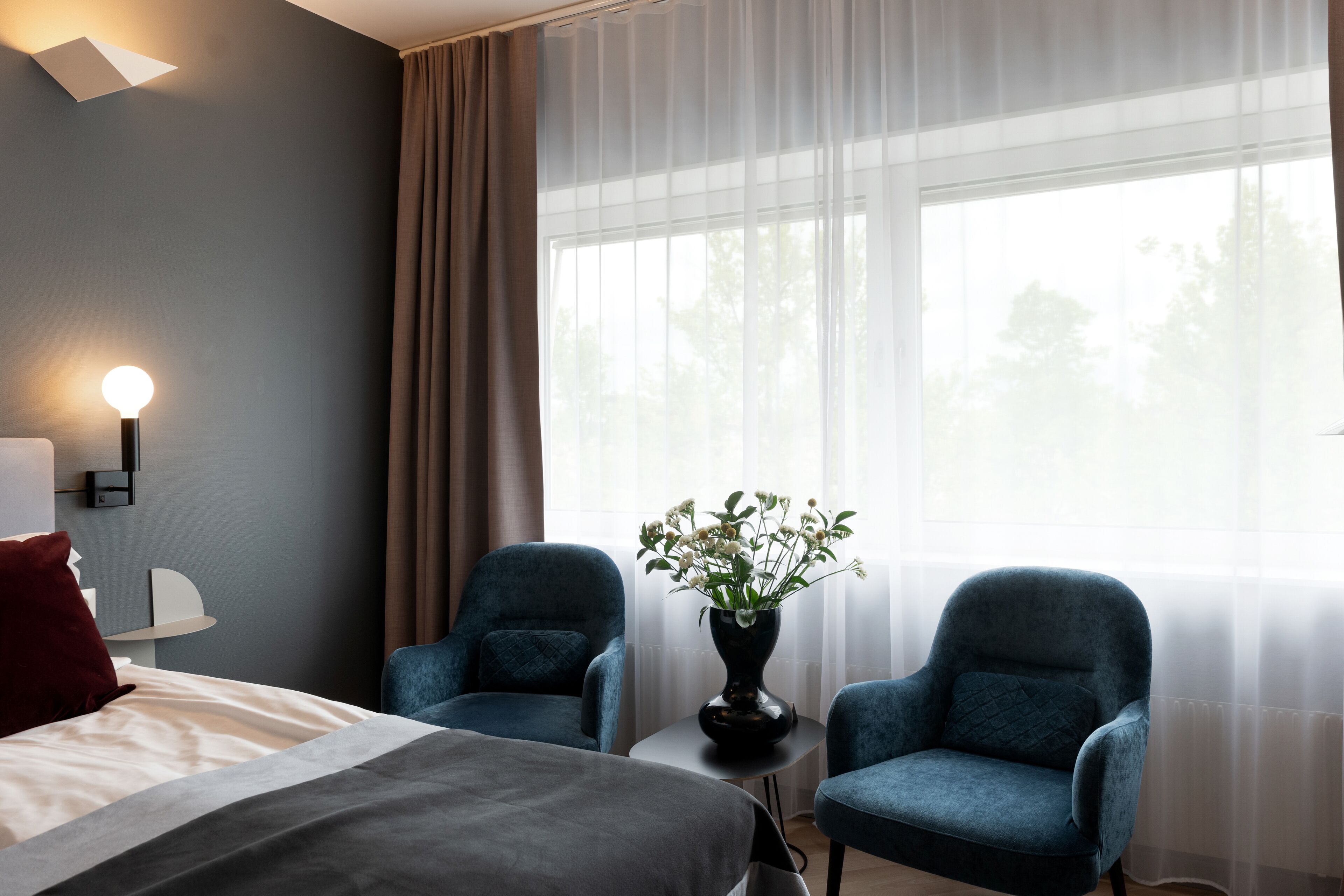 A hotel room with a bed, two armchairs, a small table with a vase of flowers, a lamp, and a window with sheer curtains.