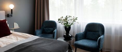 A hotel room with a bed, two armchairs, a small table with a vase of flowers, a lamp, and a window with sheer curtains.