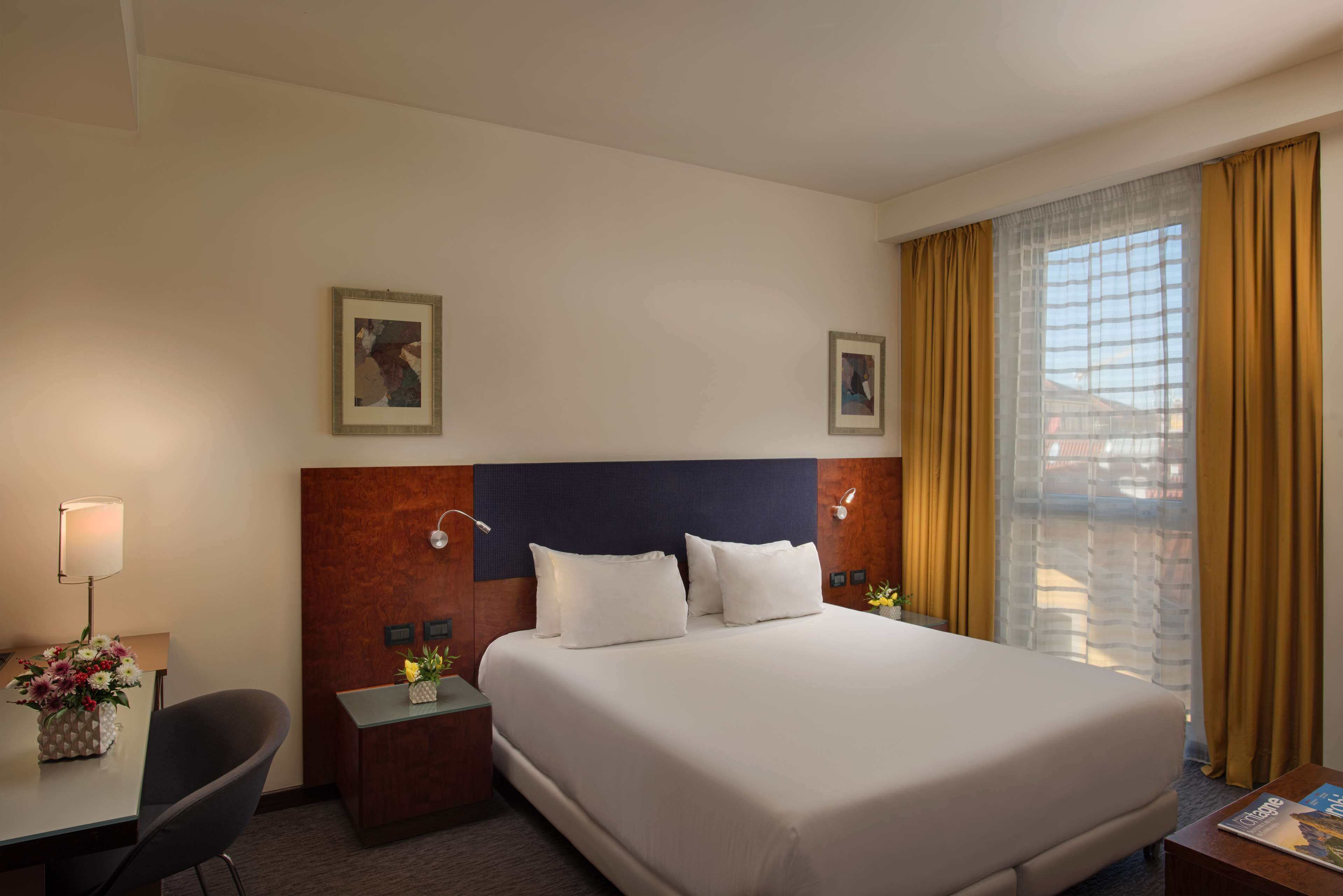 A hotel room with a large bed, a desk, a chair, a nightstand, and a window with curtains.