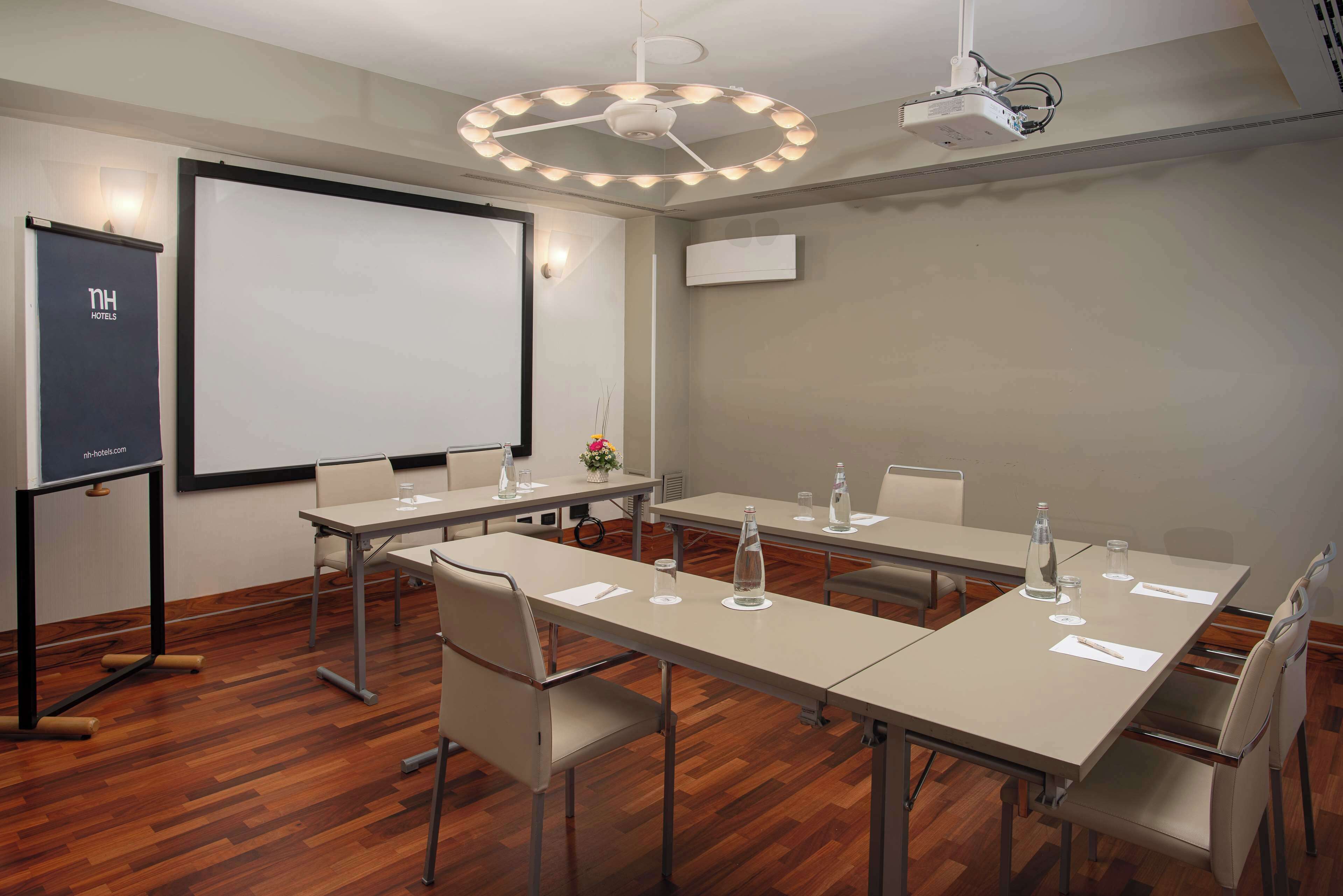 Una sala de conferencias con una gran pantalla de proyección, una pizarra y un proyector. Varias mesas con sillas y botellas de agua.
